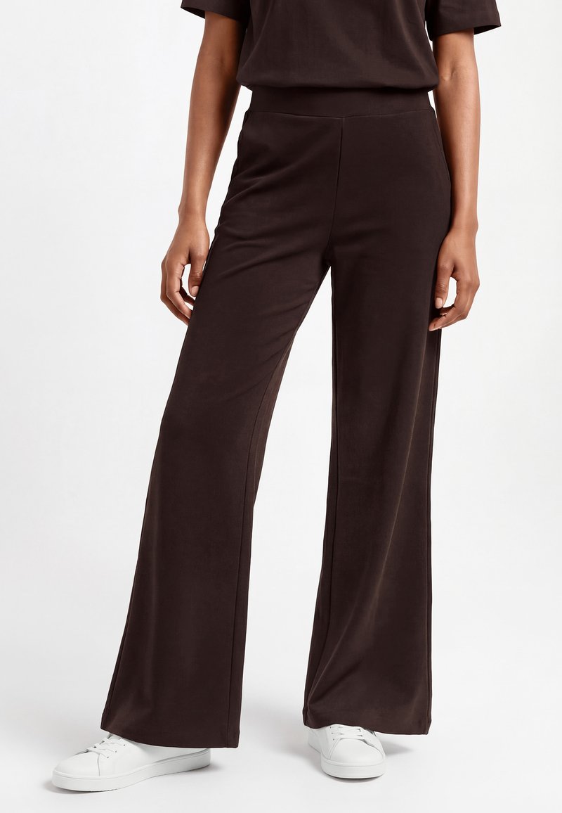 Kobieta ubrana w luźne, szerokie spodnie w ciemnym brązowym kolorze oraz białe trampki, stojąca na tle jednolitego jasnego tła.