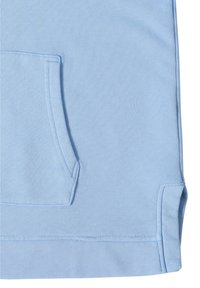 Primo piano di un tessuto azzurro chiaro che mostra una tasca a canguro con cuciture visibili e uno spacco laterale all'orlo inferiore.