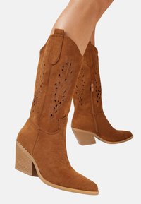 Bottes de cowboy en suède beige avec des découpes, bout pointu et talon bloc. Zip sur le côté pour un enfilage facile. Texture lisse avec une finition mate.