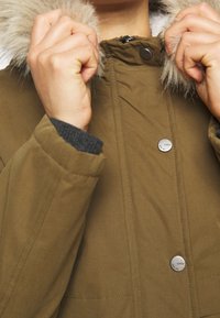 Olive green jacket with a fur-lined hood, featuring snap buttons, a quilted texture, and knit cuff detail. Hands grasping the collar.