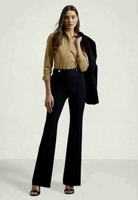 Moteris vilki smėlio spalvos marškinėlius su mygtukais, juodas aukšto liemens kelnes ir juodą švarką per petį, stovinti prieš paprastą foną.