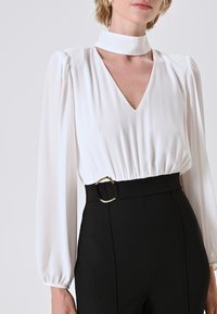 Camicia bianca con scollatura a V profonda e maniche lunghe a sbuffo, arricciata in vita. Presenta una cintura nera con una fibbia dorata.