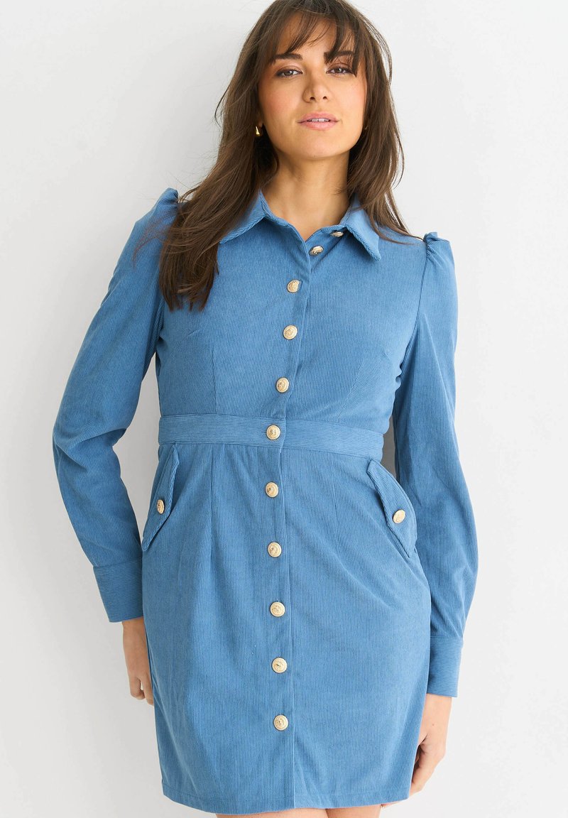 Blue corduroy shirt dress with long sleeves, front button closure, two side pockets, and a belted waist. Features a classic collar and textured fabric.