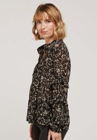 Blouse noire à fleurs avec des manches volantes, dotée d'un col boutonné. Tissu léger et transparent orné de petites fleurs blanches et beiges.