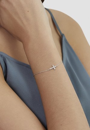 Zarter Silberkettenarmband mit einem kleinen, mit Diamanten besetzten Kreuz, das an einem Handgelenk getragen wird, vor einem blauen Satinschulterfreies Top.