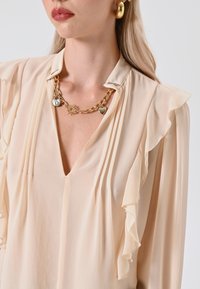 Blusa beige con maniche a ruffle, scollatura a V profonda e pieghe sottili. Accessoriata con una collana a catena dorata con ciondoli e accenti a forma di cuore.