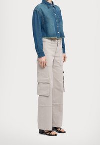 Camicia di jeans tagliata con chiusura a bottoni e orlo sfilacciato, abbinata a pantaloni cargo beige con ampie tasche e una vestibilità comoda. Sandali neri.
