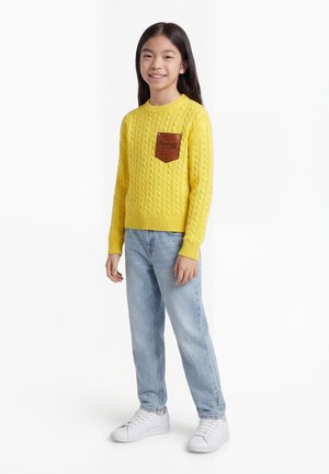 Ragazza in piedi, indossa un maglione giallo a trecce con una toppa marrone, jeans azzurri chiari e sneakers bianche, sorride alla macchina fotografica.