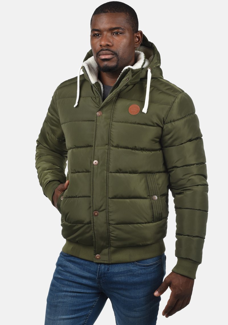 Padded green jacket with a fur-lined hood, contrasting beige accents, button closures, and elastic cuffs; featuring a logo patch on the chest.