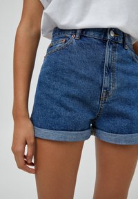Shorts de denim en azul medio con cintura alta, puños enrollados y detalles de costura sutiles. Presenta un diseño clásico de cinco bolsillos.