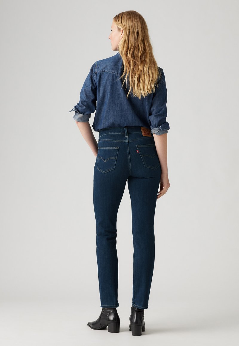 Jeans en denim bleu foncé avec une taille haute, une coupe slim et des coutures subtiles. Accompagné d'une chemise en denim foncé, vue de dos.