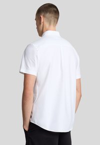 Camicia bianca a maniche corte con colletto a punta, pinces sul retro e orlo curvo. Il tessuto appare liscio e leggero.