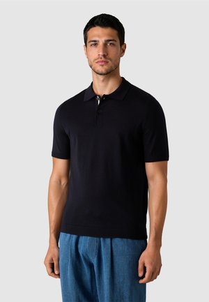 Mand iført en sort poloshirt med korte ærmer og løse blå bukser, der står mod en ensfarvet lysegrå baggrund.