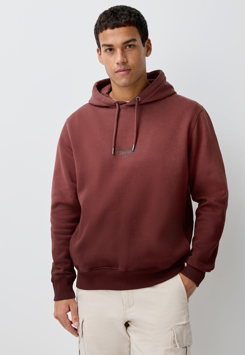 Sweat à capuche bordeaux en tissu doux, avec une poche kangourou à l'avant, une capuche à cordon de serrage et un petit logo sur la poitrine. Texture lisse, coupe décontractée.