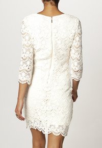 Spetsklänning i vitt, med trekvartslånga ärmar, scalloped fåll och en synlig bakdragkedja. Invecklat blommönster genom hela klänningen.