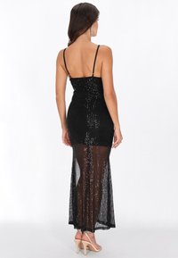 Vestido negro de lentejuelas con un corsé ajustado y espalda descubierta, que presenta una sección inferior transparente. Tirantes delgados y una silueta larga y fluida.