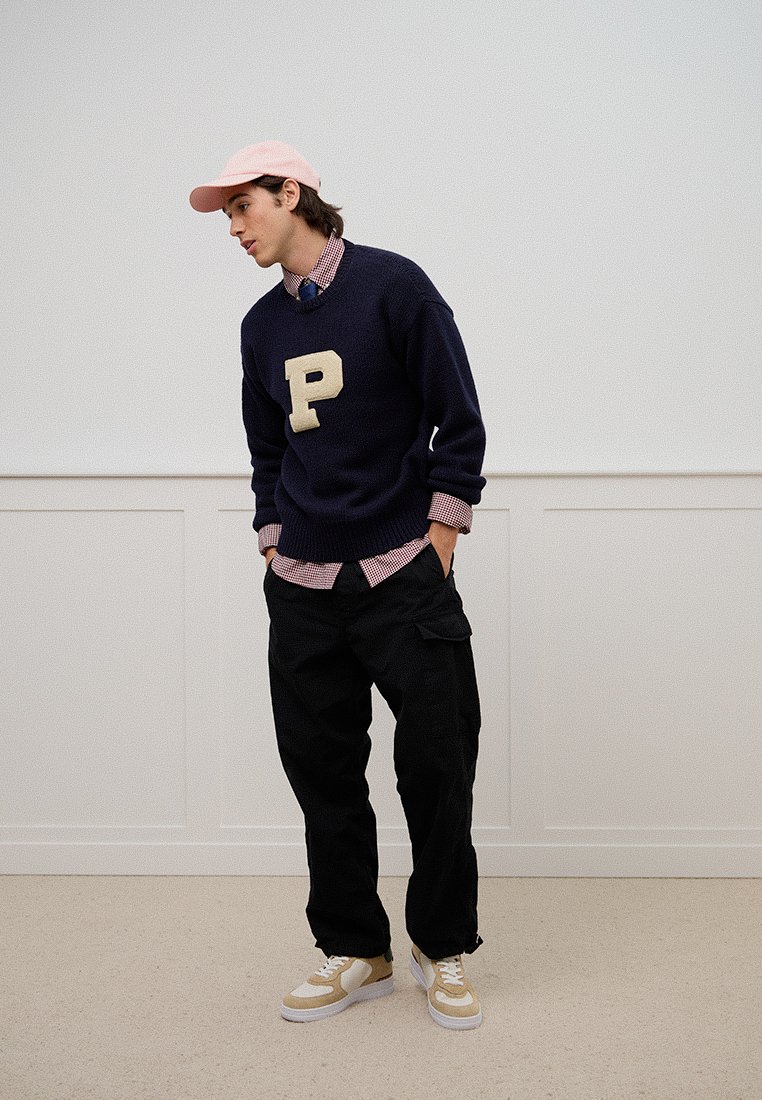 Dunkelblauer Strickpullover mit einem beigen "P"-Logo, über einem rosa karierten Hemd getragen, kombiniert mit schwarzen Cargo-Hosen und cremefarbenen Sneakers. Rote Kappe auf dem Kopf.