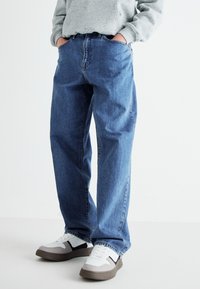 Jeans de mezclilla azul con un corte relajado, que cuentan con una cintura alta, cinco bolsillos y un ligero desgaste. Combinados con zapatillas blancas y una sudadera gris.