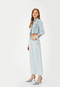 Chaqueta de mezclilla recortada en azul claro con mangas largas, combinada con jeans de pierna ancha de talle alto y tacones azules de punta con detalles de hebilla.