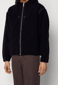 Schwarzer Fleece-Hoodie mit Reißverschluss vorne, verstellbarem Kordelzug an der Kapuze und schwarzen Seitenteilen; verfügt über eine strukturierte Stoffoberfläche und eine lockere Passform.