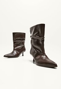 Botine din piele maro, cu un design căzut, vârfuri ascuțite și tocuri mici. Au o finisare texturată care amintește de pielea de șarpe.