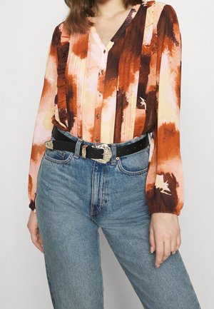Mehrfarbige Bluse mit orangefarbenen, braunen und pfirsichfarbenen abstrakten Mustern; lange Ärmel, Knopfverschluss-Design und strukturierter Stoff, kombiniert mit blauen Jeans.