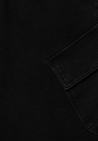 Tissu en denim noir avec une texture lisse et des détails de couture visibles. Un rabat de poche est partiellement visible, renforçant le design.