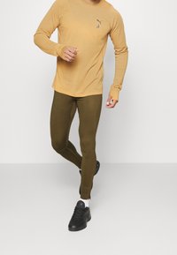 Hořčicová dlouhá košile s malým černým logem, zkombinovaná s olivově zelenými legínami a černými sportovními botami. Hladký textura materiálu.