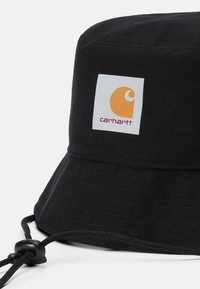 Sort bøttehat lavet af holdbart stof har en rund krone og en bred, nedadgående skygge. Inkluderer et logo-patch og justerbar snor.