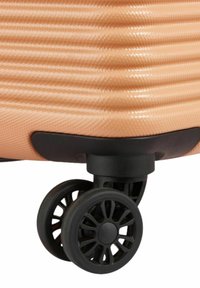 Valigia rigida color pesco con superficie testurizzata, dotata di una ruota nera con design a raggi per la mobilità. Base nera accentuata.