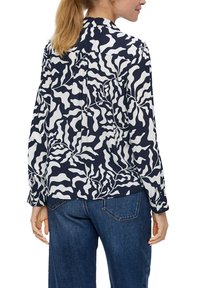 Kvinna med blont hästsvans som bär en långärmad marinblå och vit abstrakt mönstrad skjorta och blå jeans, sedd bakifrån.