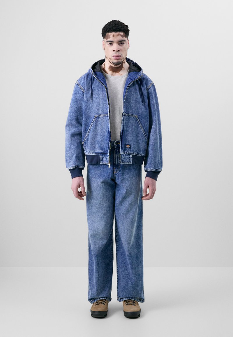 Giubbotto di denim con cappuccio, blu, con tasche a marsupio. T-shirt grigia sotto, abbinata a jeans blu a gamba larga e stivali color cammello.