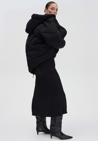 Doudoune noire avec une grande capuche, motif en losanges, associée à une jupe noire ajustée et des bottes à talons aiguilles jusqu'au genou.