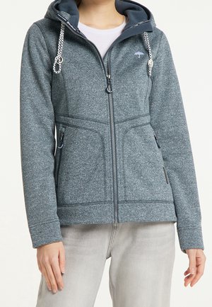 Kvinde iført en grå hættetrøje med frontlynlås, snoretræk og lynlåslommer i siderne, kombineret med lyse beige bukser.