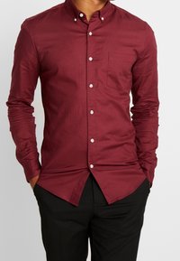 Muž v zapínacím burgundském dlouhém rukávu s kapsou a černých kalhotách, s rukama v kapsách, na jednobarevném pozadí.