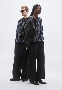 Dve manekenki nosita ohlapne pletene puloverje v črnih in modrih vzorcih, kombinirane s širokimi črnimi hlačami. Ena drži črno usnjeno jakno.