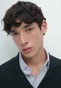 Młoda osoba dorosła o kręconych ciemnych włosach ma na sobie czarny sweter z prążkowanym kołnierzem na jasnoniebieską koszulę na guziki. Tło jest neutralne.