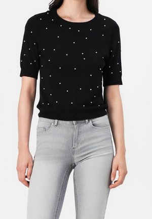 Sort kortærmet sweater med små hvide perledekorationer, båret med lysegrå højttaljede jeans.