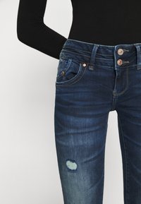 Mörkblå denimjeans med en slitning vid knäet och en fransad hål, dubbel knappstängning och klassisk femfickdesign.