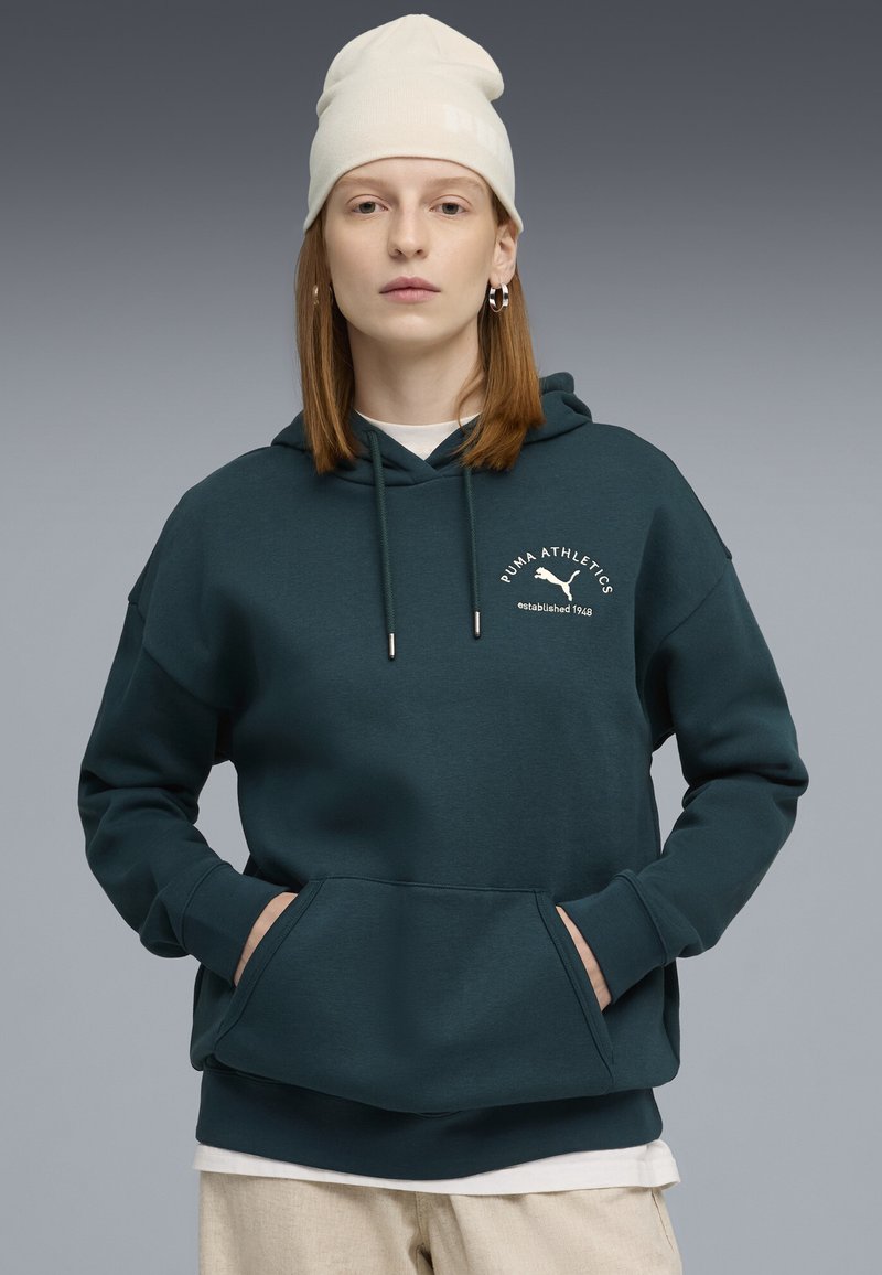 Felpa di colore verde scuro con tasca a marsupio, cappuccio regolabile con cordino e dettagli del logo bianchi. Indossata con una maglietta bianca sotto. Berretto crema indossato.