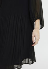 Schwarzes plissiertes Kleid aus transparenter Stoff, mit langen Ärmeln und schwingender Silhouette, mit einer subtilen Textur und schichtartigem Erscheinungsbild.