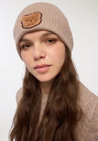 Beżowa dziana czapka z prążkowanym wzorem i brązową łatą w kształcie niedźwiedzia z przodu; noszona z luźnymi, falowanymi brązowymi włosami.