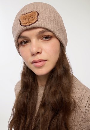 Beżowa dziana czapka z prążkowanym wzorem i brązową łatą w kształcie niedźwiedzia z przodu; noszona z luźnymi, falowanymi brązowymi włosami.