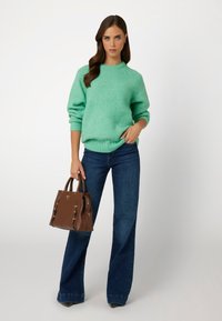 Maglione strutturato verde chiaro con scollo rotondo, abbinato a jeans a vita alta blu scuro svasati. Tiene una borsa rigida marrone.