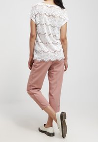 Top in pizzo bianco con motivo a scallop, abbinato a pantaloni rosa pallido e scarpe bianche con tacco basso. Texture morbida e vestibilità rilassata.