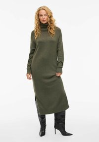 Robe tricot olive avec un col roulé haut, des manches longues, des poignets côtelés et des fentes sur les côtés, associée à des bottes noires à talons montant jusqu'au genou.