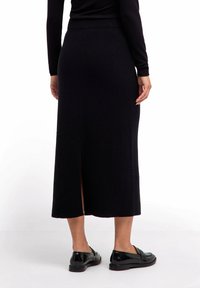 Frau in einem schwarzen gerippten Midikleid mit einem Schlitz auf der Rückseite, schwarzem Langarmoberteil und schwarzen Leder-Loafers, die auf einem weißen Boden steht.