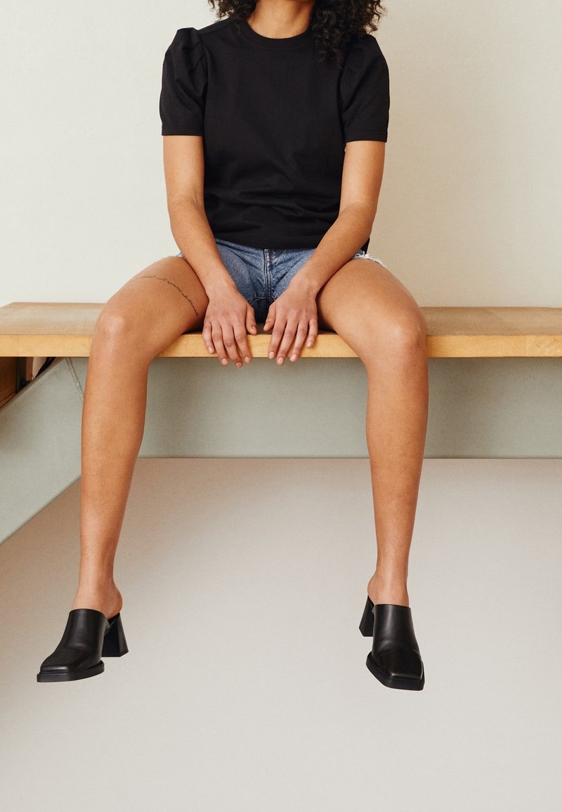 Top negro con mangas abullonadas, pantalones cortos de mezclilla y mules de tacón bloque negro. Los mules tienen un acabado de cuero suave y un diseño de espalda abierta.