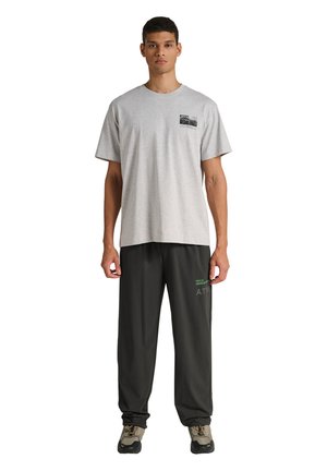 Ung mand, der står, iført en lysegrå T-shirt med et lille brystmotiv og løse sorte bukser med grøn og grå tekst på låret.