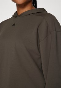 Gros plan d'une personne portant un sweat à capuche vert olive avec un petit logo noir sur la poitrine et une coiffure tressée.